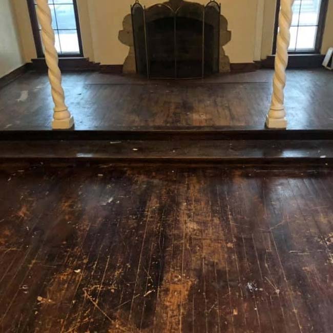 Worn hardwood floor with scratches, two twisted columns, and a stone fireplace—ideal for refinishing.